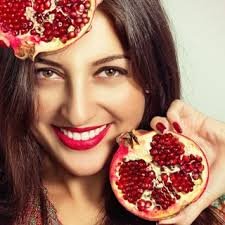 امرأة تبتسم وتمسك بثمرة رمان مفتوحة تظهر حباتها الحمراء اللامعة، مع وضع قطعة أخرى من الرمان على رأسها، مما يعكس جمالية الفاكهة وفوائدها الصحية